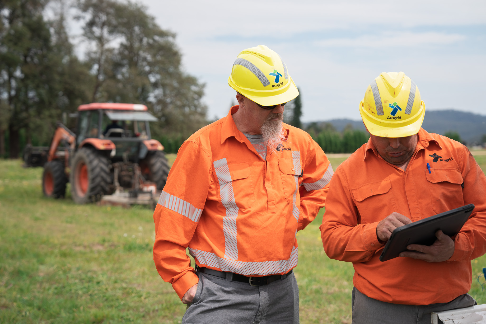 Two ausgrid field workiers with an ipad in rural nsw. A tractor is parked behind them in a field.